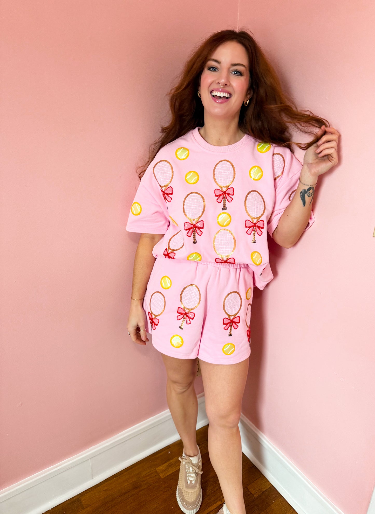 A person wearing a pink top with a tennis racket and bow pattern and shorts, standing against a pink background.