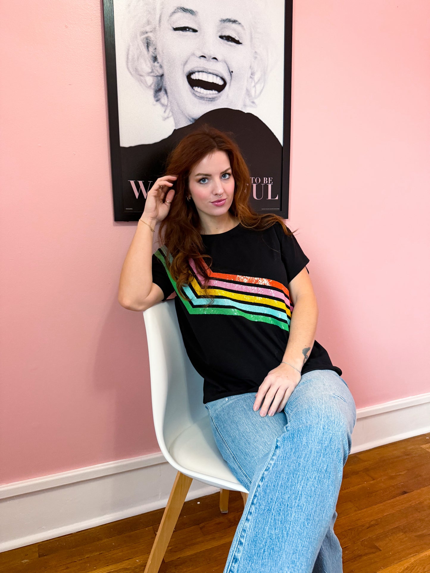 A person wearing a black t-shirt with diagonal rainbow stripes, sitting in a chair with a pink background and a poster of Marilyn Monroe in the background.
