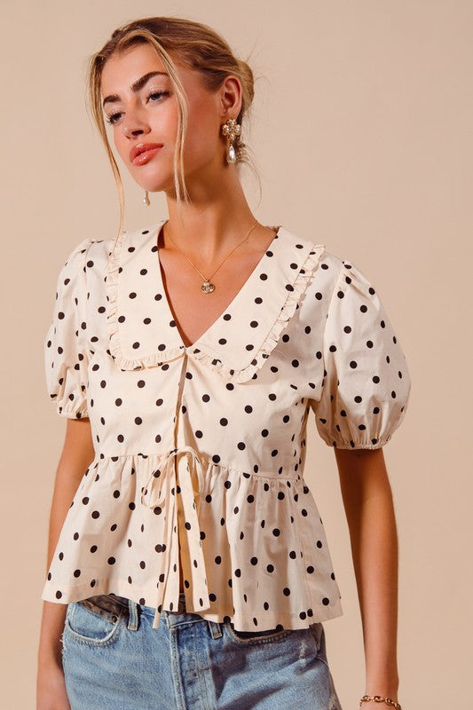 A woman wearing a beige vintage-style blouse with black polka dots, featuring a Peter Pan collar and tie-front bows.