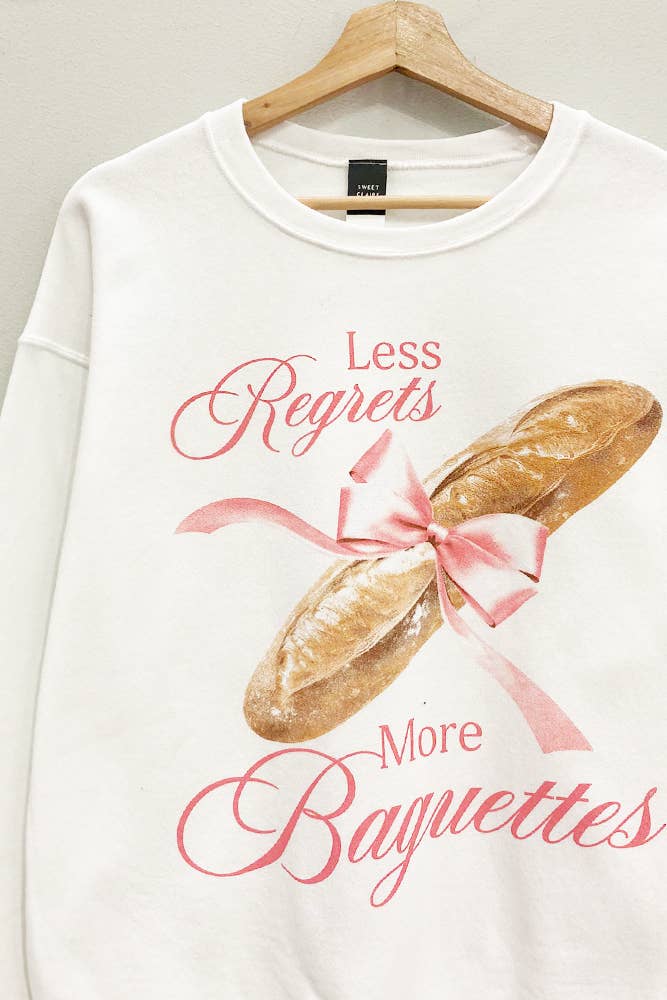 A woman wearing a white sweatshirt with the text 'Less Regrets More Baguettes' printed on the front.
